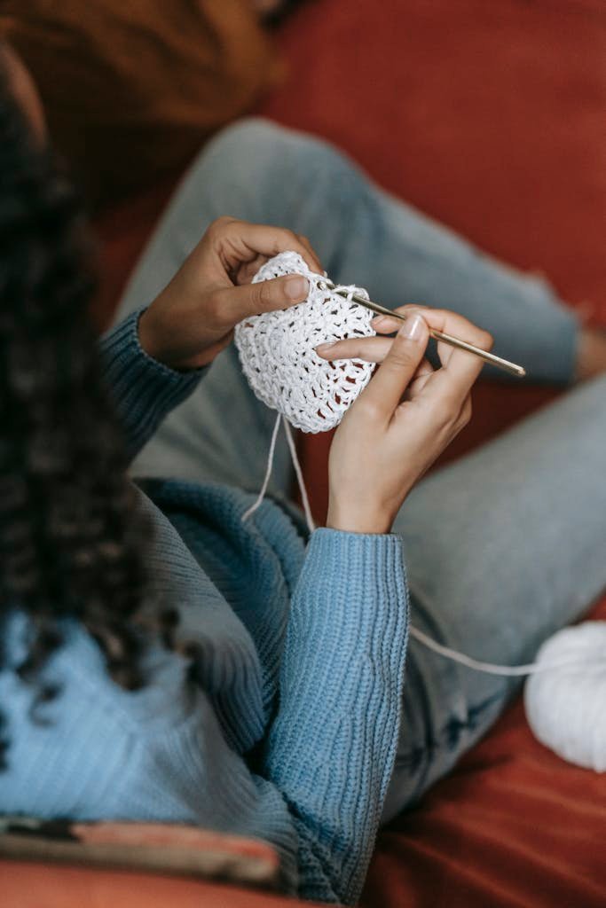 Close-up of a woman crocheting with white yarn on a red sofa, emphasizing DIY and textile craftsmanship.